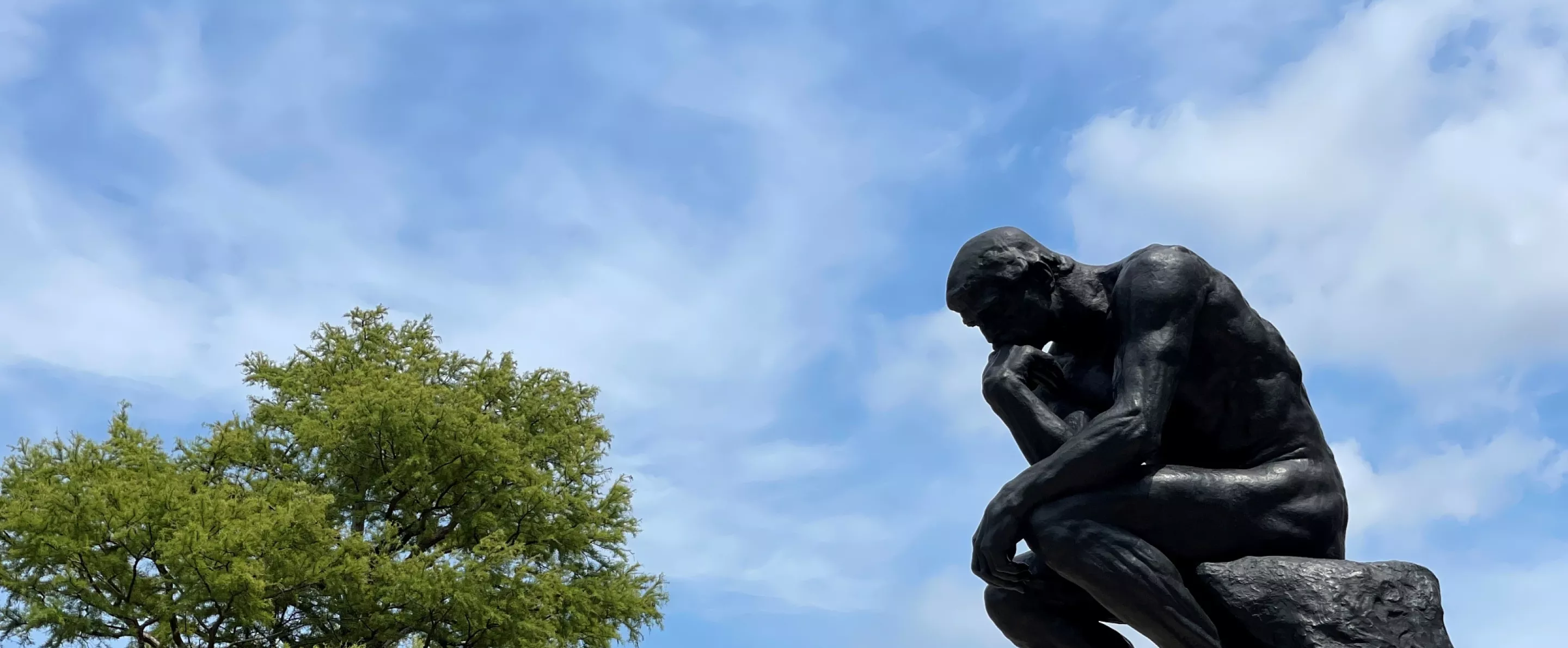 UofL's thinker statue from the side with the sky in the background. Trees on campus can be seen in the background as well as a lamp post with a UofL banner.