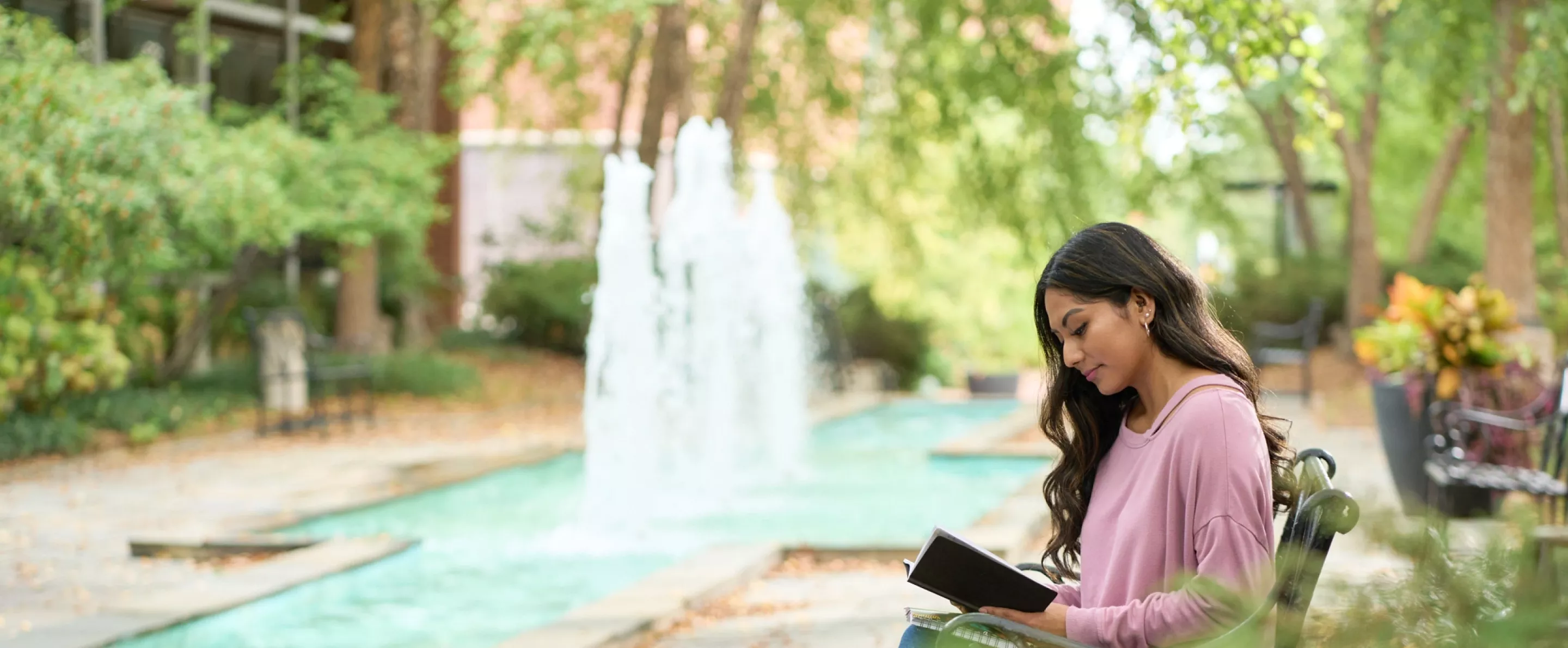 Student siting outside near fountain area reading a book while sitting on a bench.