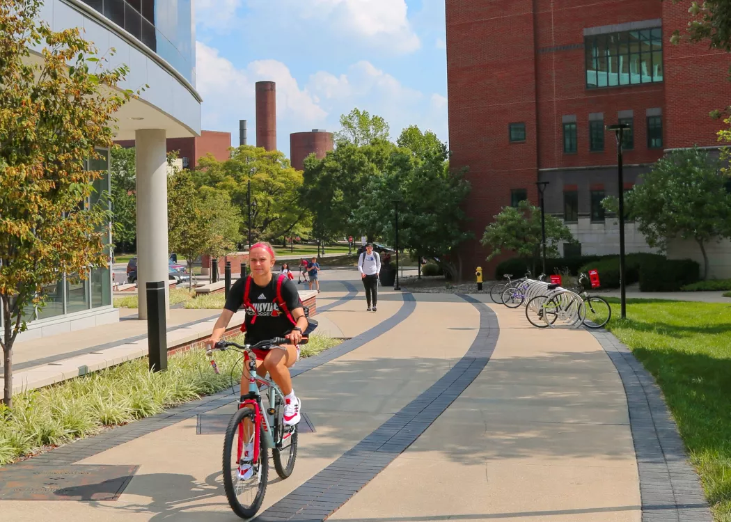 A sidewalk passing by the reflective glass Belknap Academic Building. The curving sidewalk has a student passing on a red bike wearing a black UofL T-Shirt. The day is sunny with white clouds.