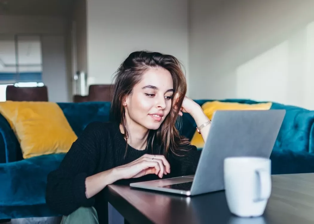 Young female student sitting on floor working on laptop at home in living room.