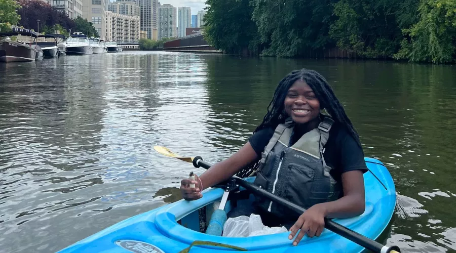 A student smiles while sitting in a bright blue kayak with the Chicago skyline in the background